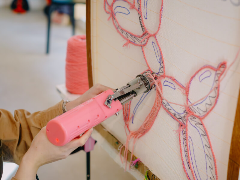 A close up of a woman making a pink balloon animal rug using a tufting gun and pink yarn